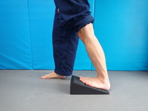 Wade dehnen – Zwillingswadenmuskel (Musculus gastrocnemius): Platziere den zu dehnenden Fuß so, dass der Fußballen gegenüber der Ferse erhöht liegt. Dafür kannst du zum Beispiel einen Wadendehner (Keil), einen halbrunden Foamroller oder einfach ein zusammengerolltes Handtuch unter dem Fußballen platzieren. Der Fuß sollte gerade nach vorne schauen. Steige nun mit dem anderen Bein nach vorne, bis zu ein Ziehen in der Wade spürst. Halte das Knie des Beins, das gedehnt wird, durchgestreckt und lass ein Gewicht auf dem Bein, das gedehnt wird. Halte die Dehnung 2 min/Bein.